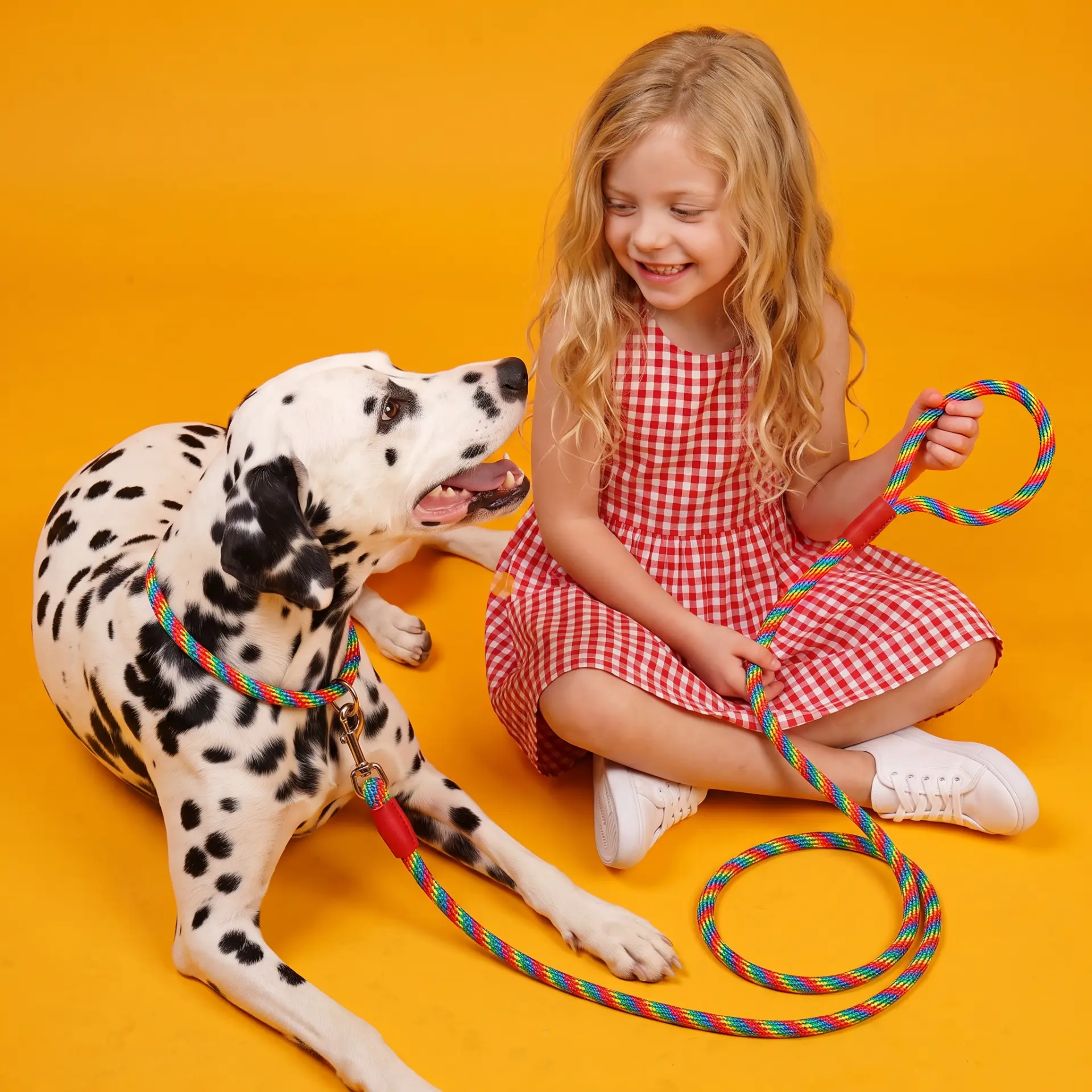 Girl sitting beside a dalmatian holding a rainbow rope leash on a yellow backdrop