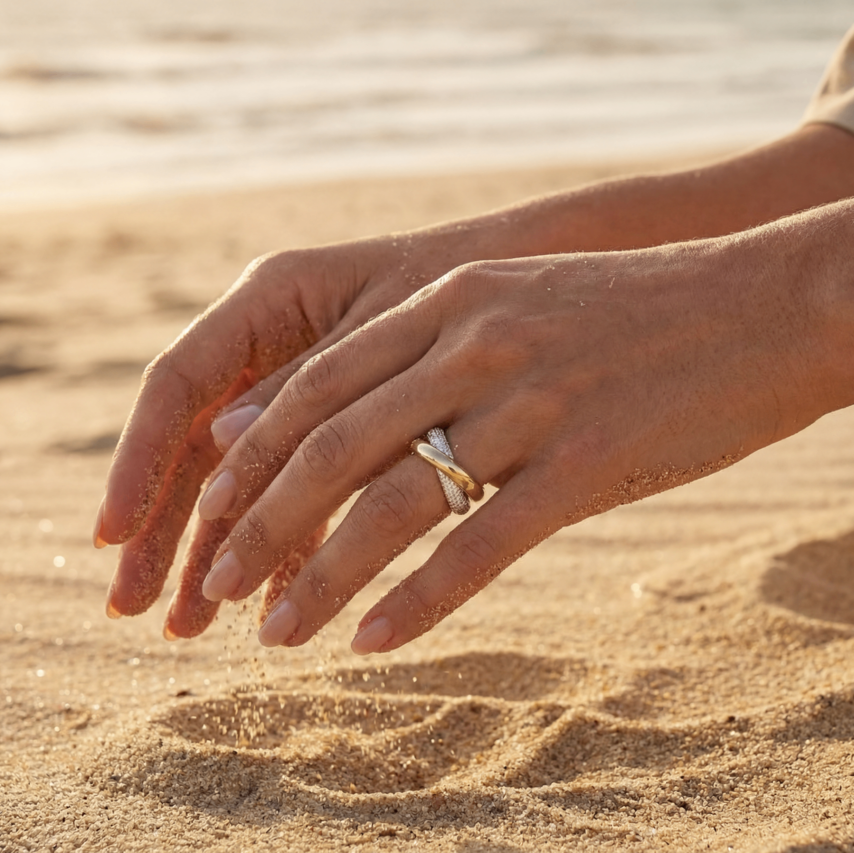 Hand running through beach sand wearing gold and diamond rings