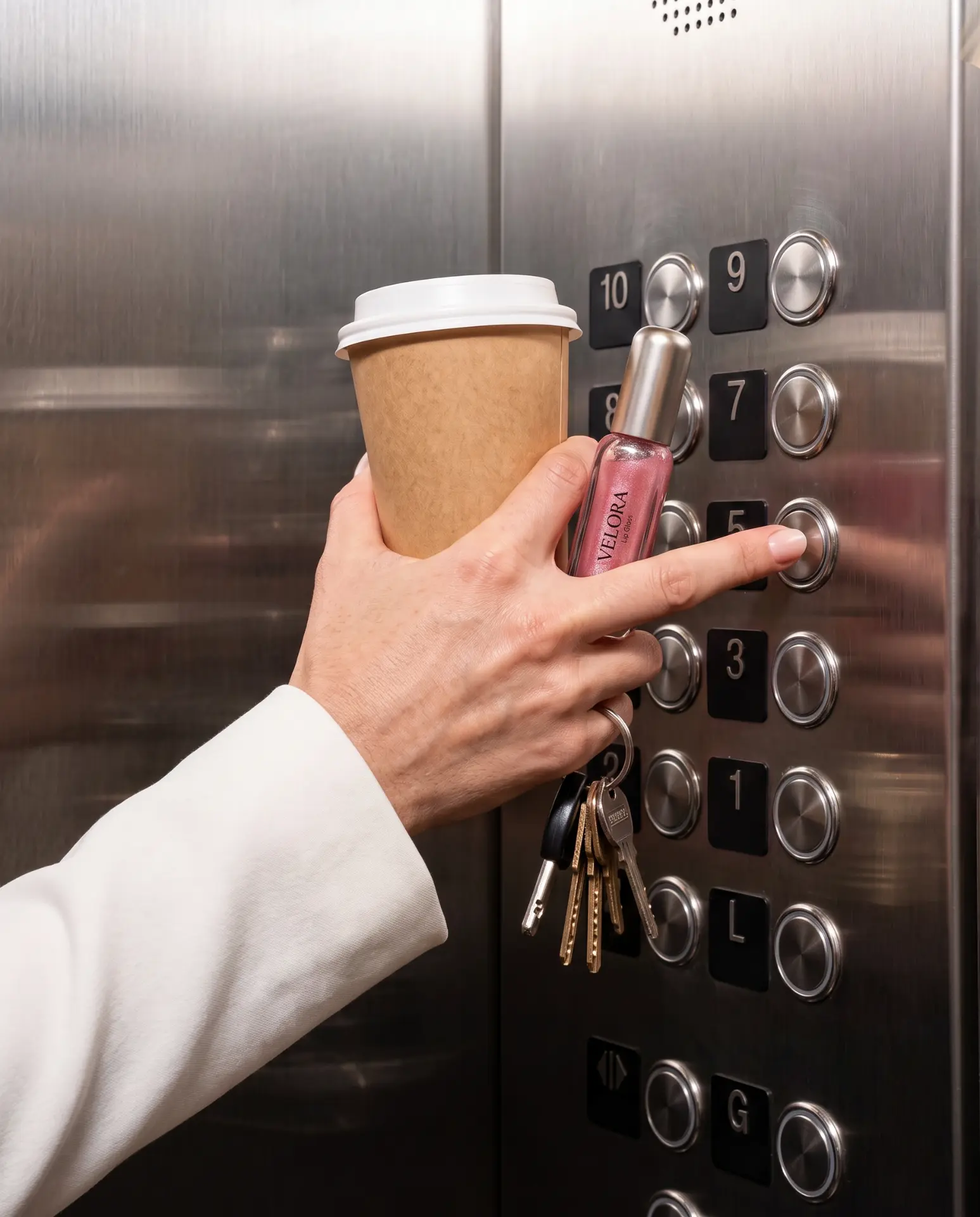 Hand holding a coffee cup, keys and a VELORA lip gloss while pressing an elevator button