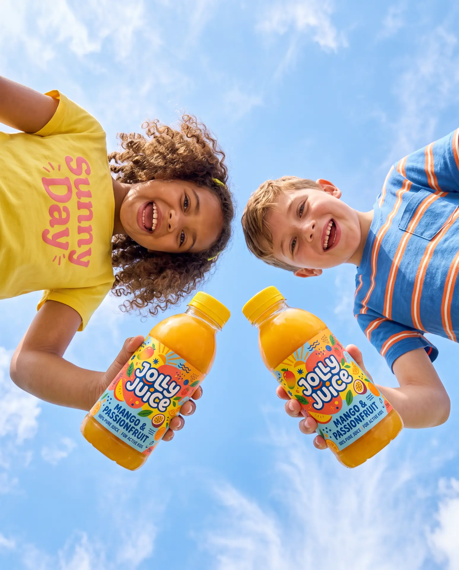 Two happy children holding Jolly Juice mango passionfruit bottles against a bright blue sky